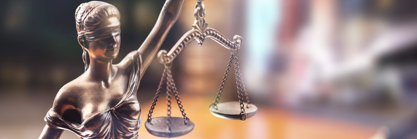 A close-up of the bronze statue of lady justice, focusing on her blindfold and balanced scales, with a blurred background suggesting a courtroom setting typically studied in criminal justice PhD programs.