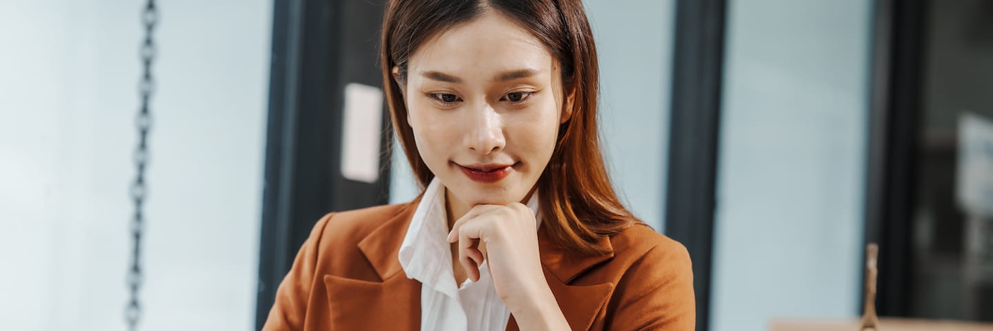 A young Asian businesswoman in a brown blazer, deep in thought about online criminal justice degrees, resting her chin on her hand while looking at papers on her desk in an office setting.