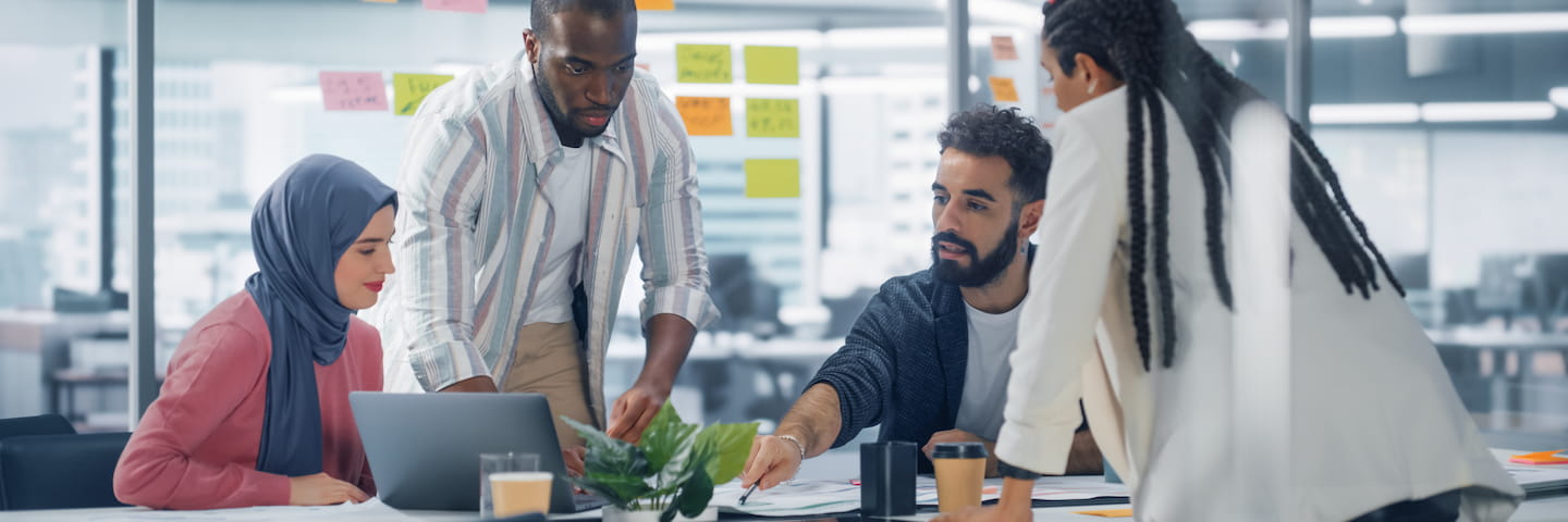 Diverse group of colleagues collaborating around a laptop in a modern office, discussing digital marketing work with sticky notes in the background.
