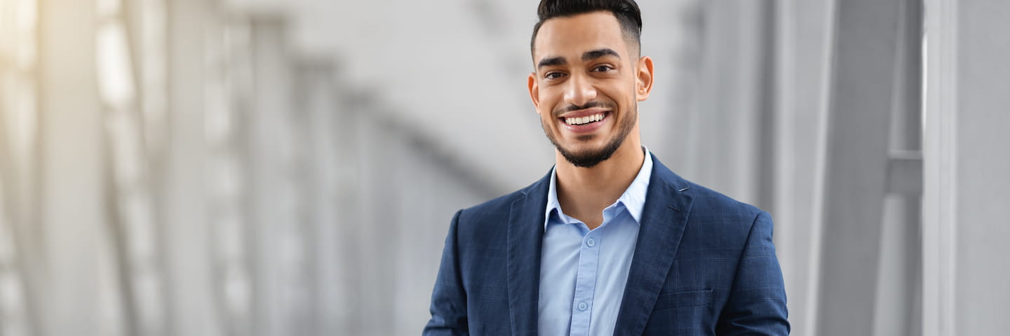 A smiling young man in a tailored blue suit stands confidently in a modern office environment, ready to discuss the best DBA programs for 2024.