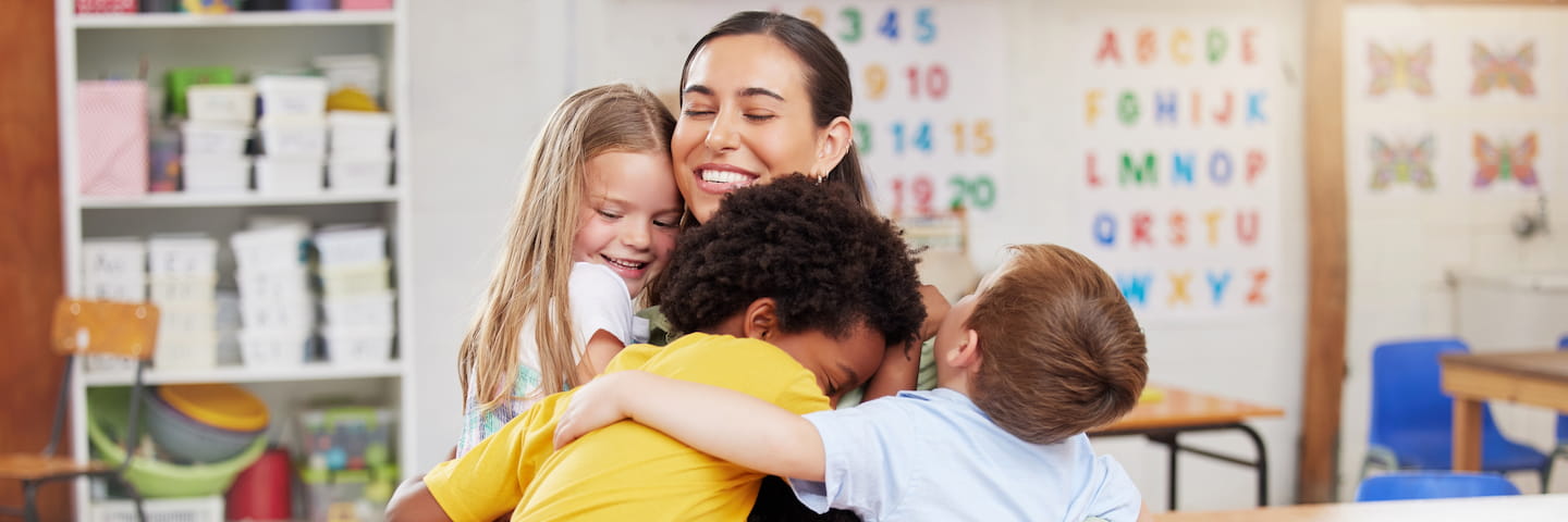 A joyful female teacher embraces three young students in a colorful classroom, surrounded by educational posters and bookshelves, promoting the best online education.