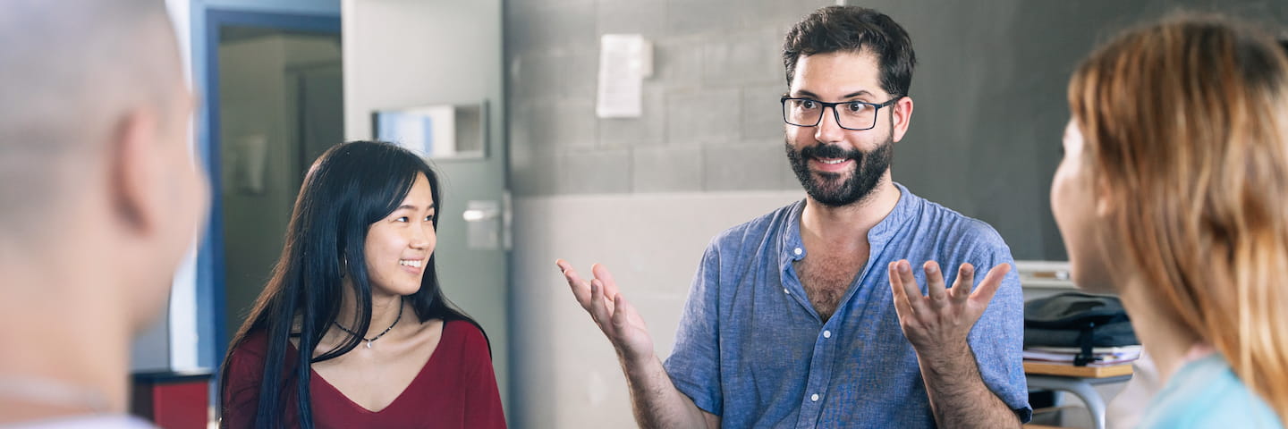 A diverse group of young adults engaging in a lively discussion about online Bachelor’s degrees in a modern office setting. A bearded man gestures animatedly while speaking to two attentive women.
