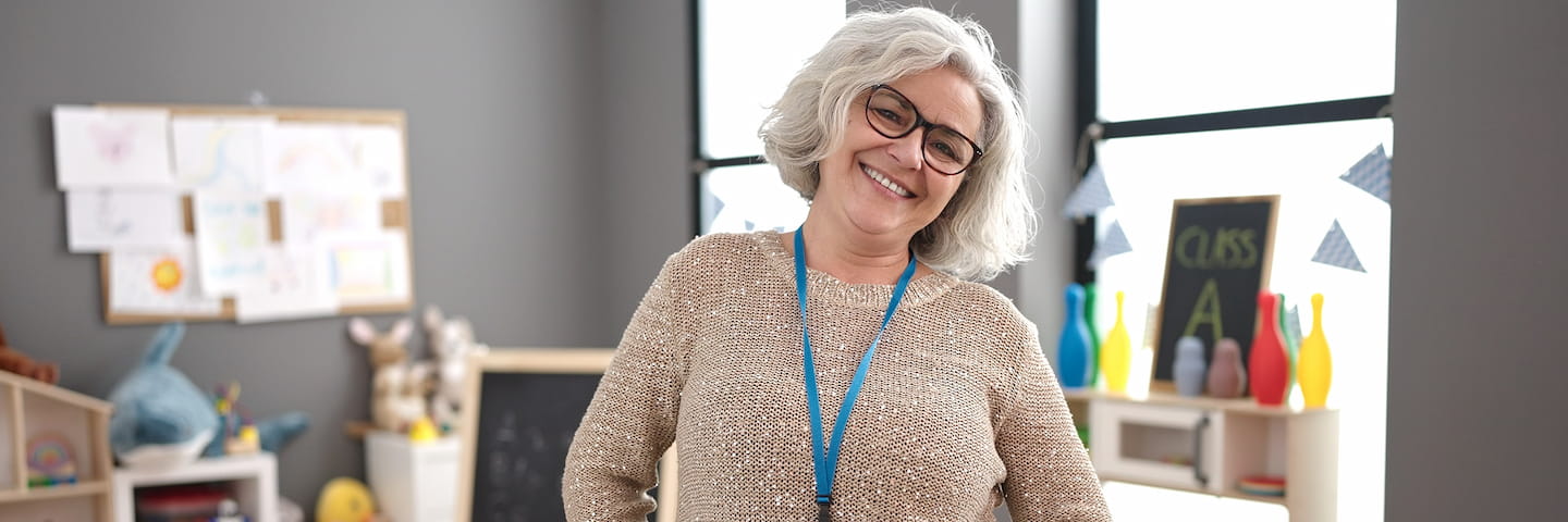 A smiling elderly woman with silver hair and glasses, wearing a beige sweater and a blue lanyard, stands in an education classroom with colorful supplies and drawings in the background.