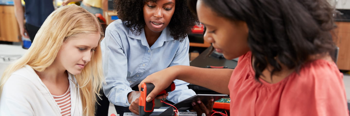 Three women, one using multimeter probes on a circuit board, discussing electronics in a workshop setting for their 2024 Online Engineering Degrees program.