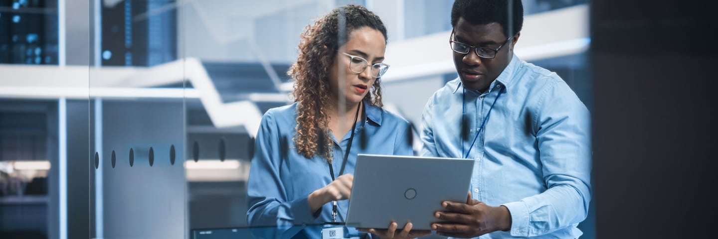 Two professionals, a woman and a man, discuss online engineering degrees over a tablet in a modern office environment, focusing intently on the screen.