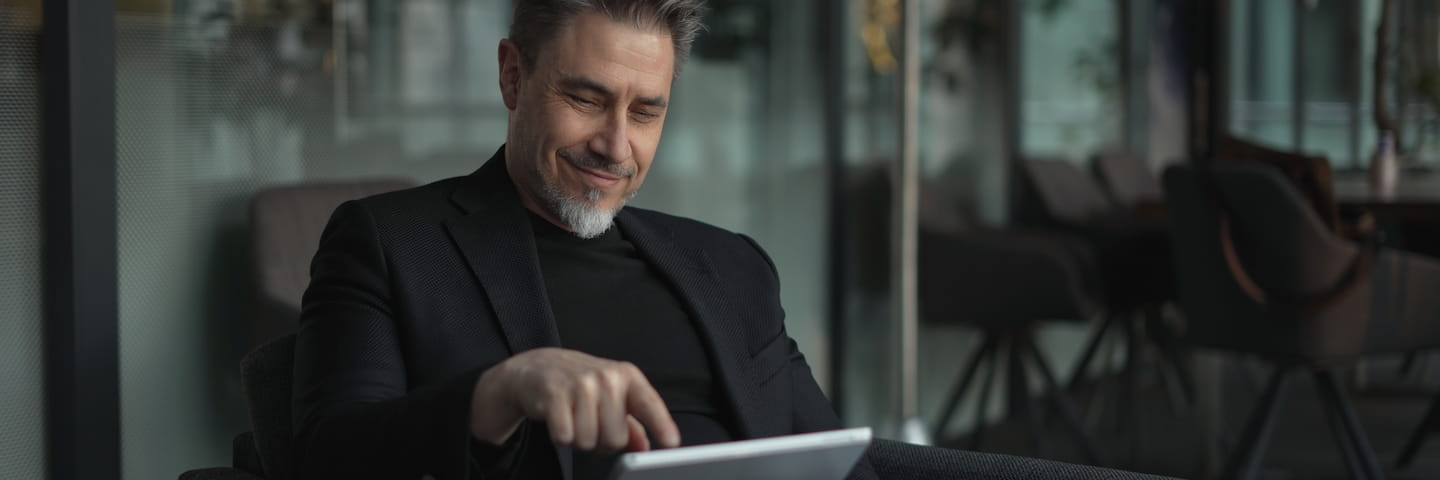 A smiling, middle-aged man with graying hair, dressed in a black blazer, uses a tablet to explore online entrepreneurship degree options in a modern office setting with large windows.