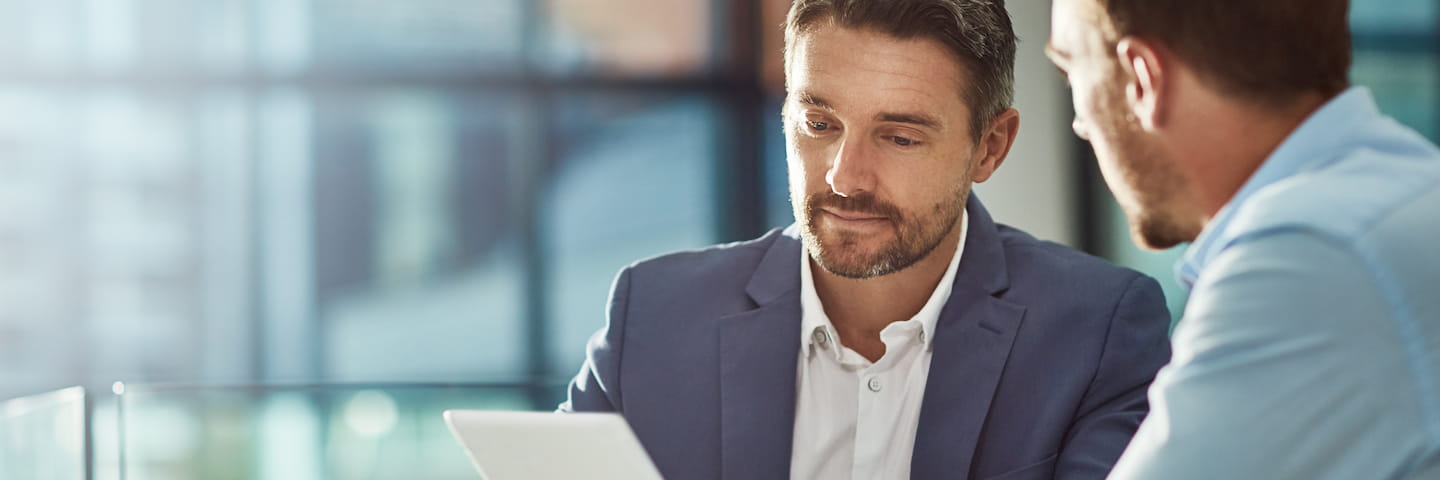 Two businessmen in a discussion about top EMBA online programs, one holding a tablet, in a brightly lit office environment with a modern glass backdrop.