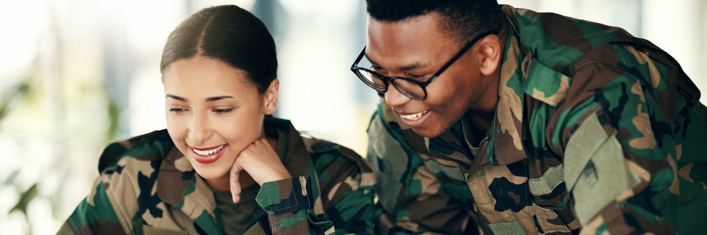 Two smiling military personnel, a man and a woman in camouflage uniforms, looking at a screen together with a light, engaging expression in an indoor setting, possibly discussing military tuition benefits.