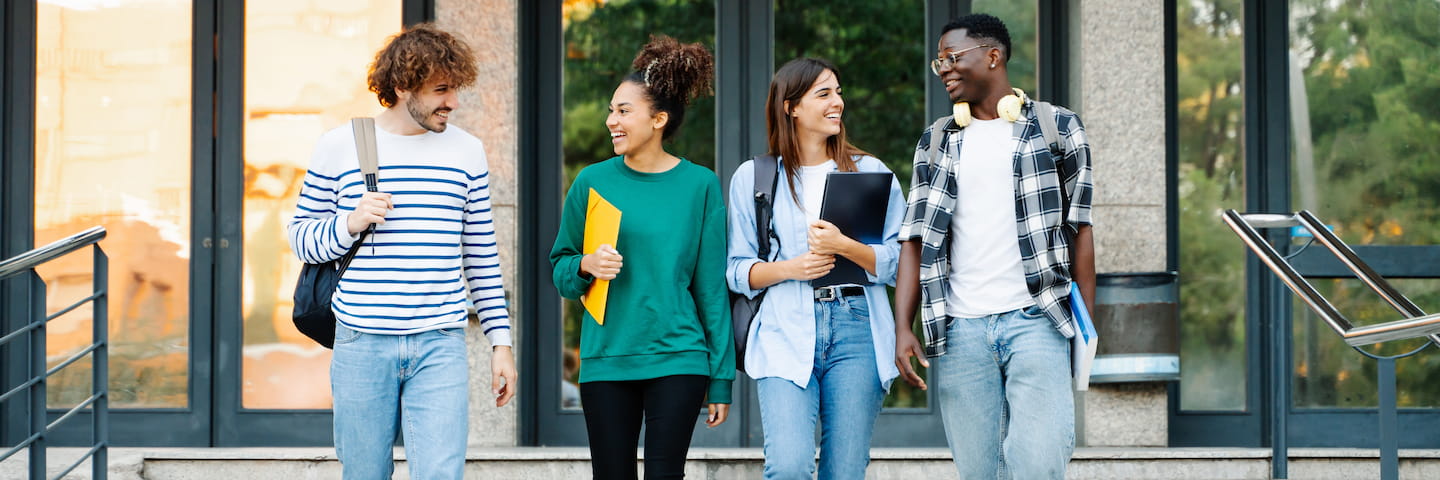 Four students of diverse backgrounds, all recipients of college scholarships, walking and chatting happily outside a university building, carrying books and backpacks.