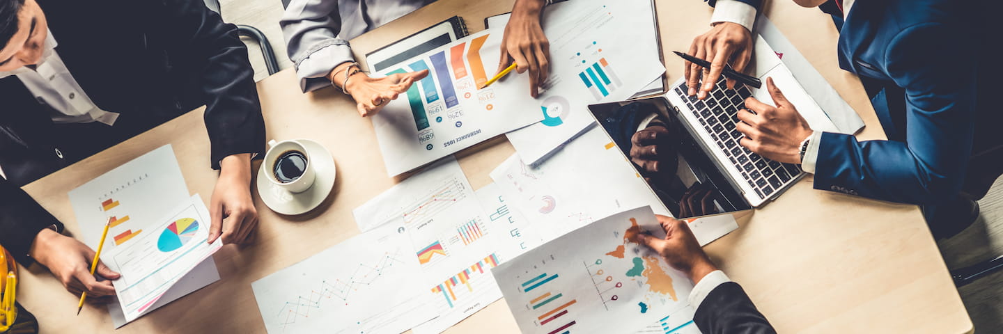 Top view of a business meeting with six professionals around a table covered with charts, a laptop, smartphone, and coffee cup, engaging in a discussion about financial aid.