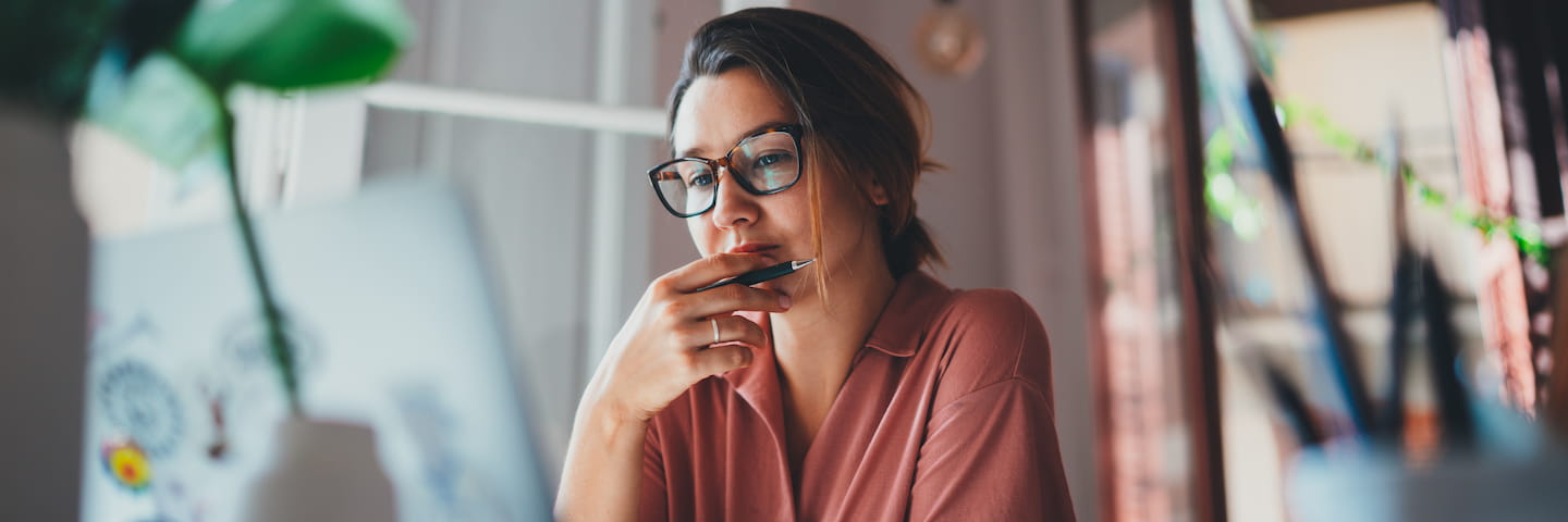 A woman wearing glasses pensively holds a pen near her mouth while looking at a computer screen, surrounded by houseplants in a cozy indoor setting, likely researching Full-Time MBA programs.
