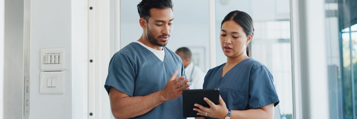 Two healthcare management professionals, a man and a woman, wearing blue scrubs, standing in a hospital corridor and focused on a digital tablet one of them is holding.