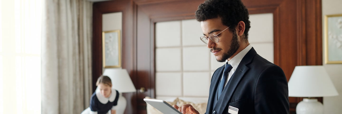A young man in a suit reviews notes on a tablet about Online Hotel Management in a well-appointed room with a woman cleaning in the background.