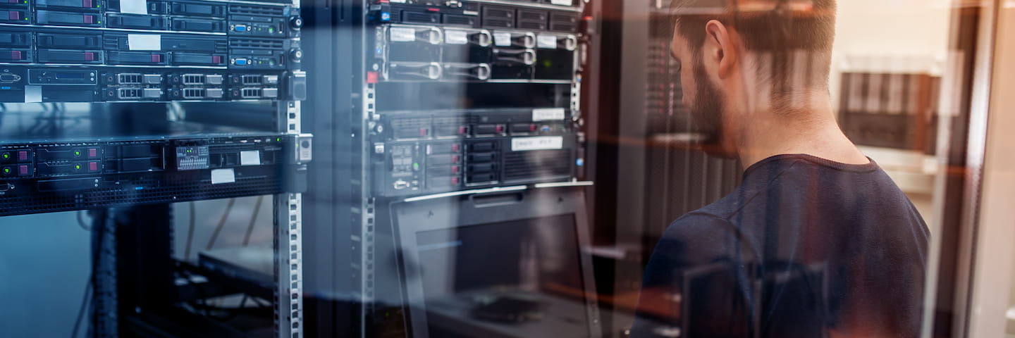 A man in a dark shirt inspects servers in a data center, observing the Information Systems equipment intently from a close distance, with a reflection visible on the glass door.