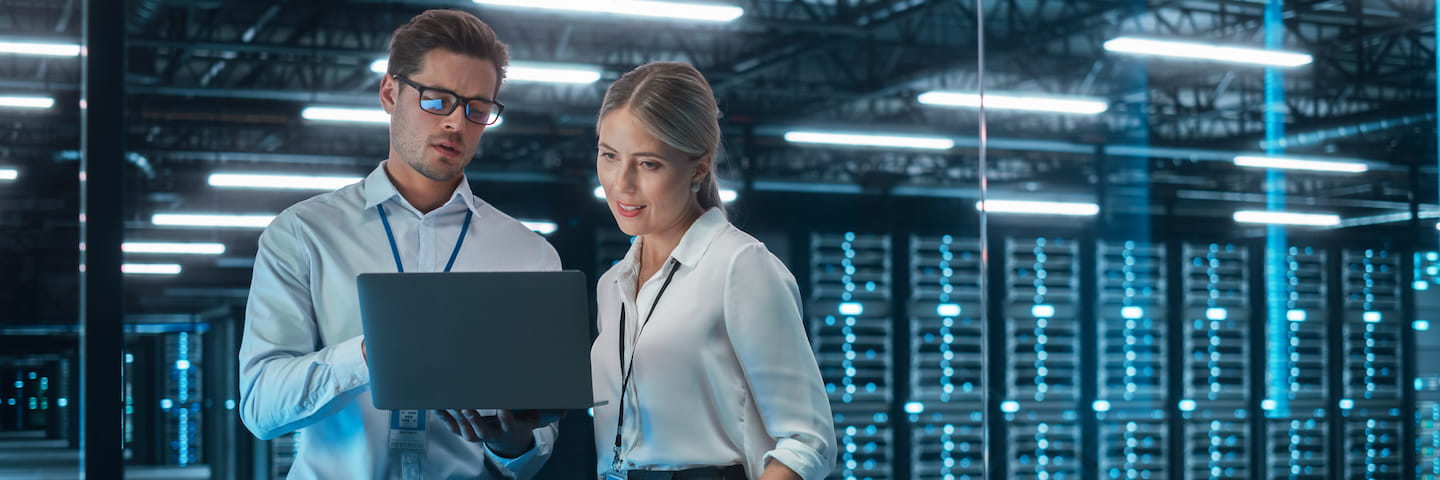 Two professionals, a man and a woman, examine a laptop together in a well-lit server room with rows of computer servers in the background while discussing Online MBA Programs IT.