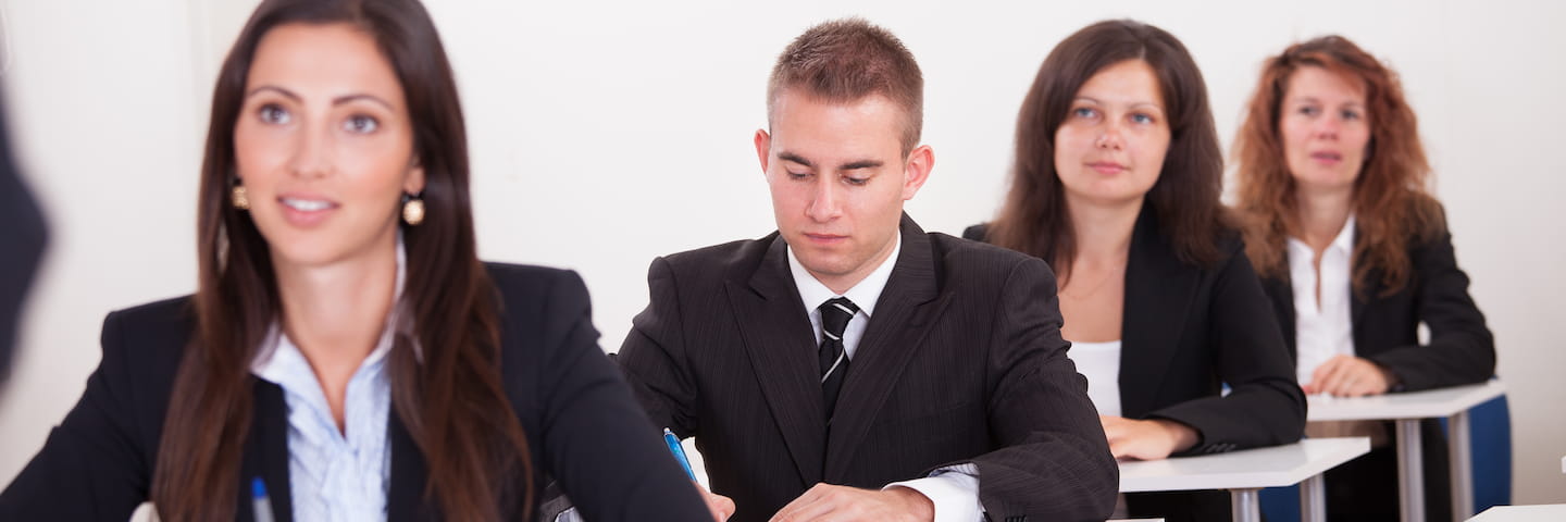 Four young professionals sit in a row at a business seminar, with one man in the foreground taking notes and three women looking attentive.