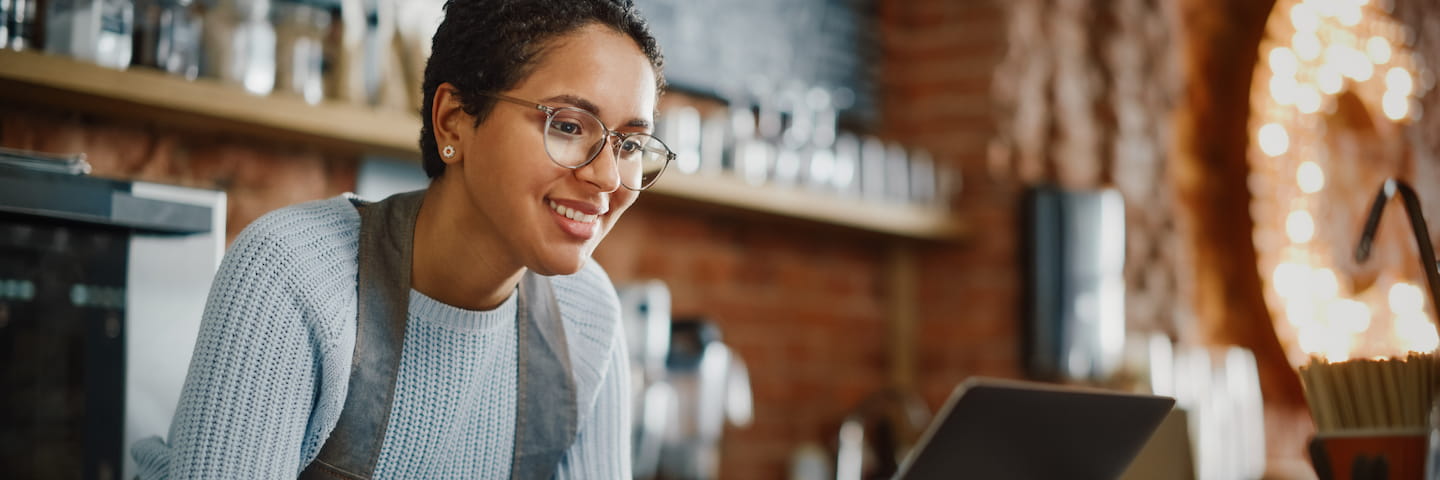 A smiling entrepreneur with glasses working on a laptop in a cozy cafe decorated with string lights and shelves full of jars.