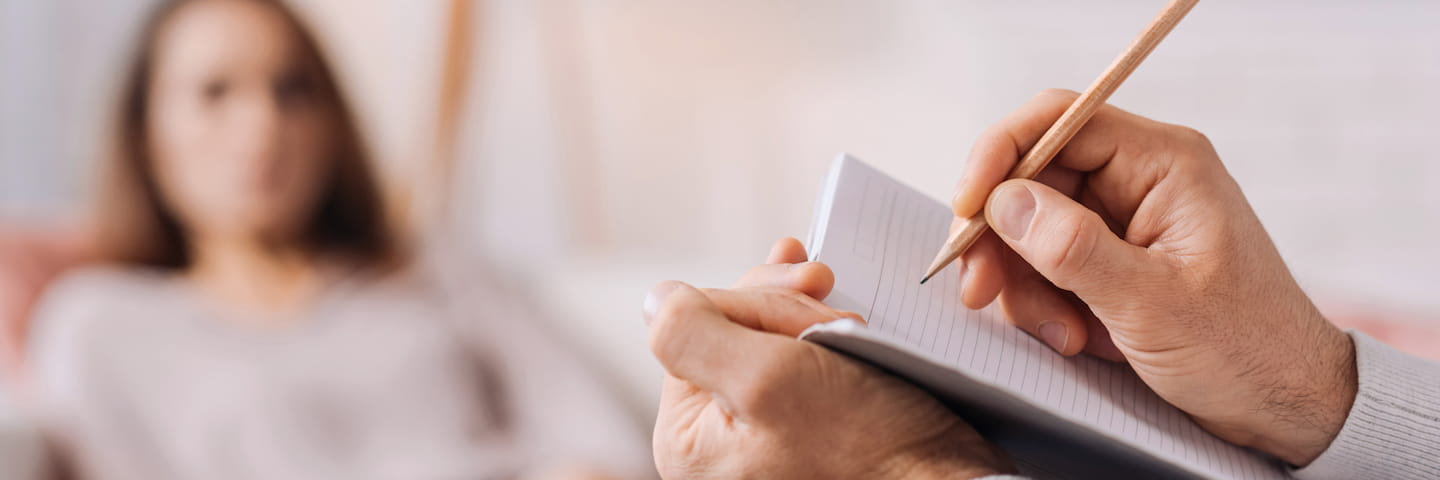 A therapist enrolled in one of the best psychology programs writes notes on a pad while a female patient sits blurred in the background during a counseling session. Focus on therapist's hands.