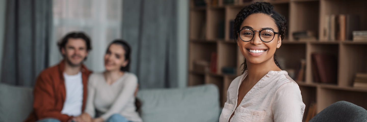 A young woman wearing glasses smiles at the camera, with a couple sitting on a couch slightly blurred in the background, inside a cozy living room. She holds a brochure for online psychology degrees.