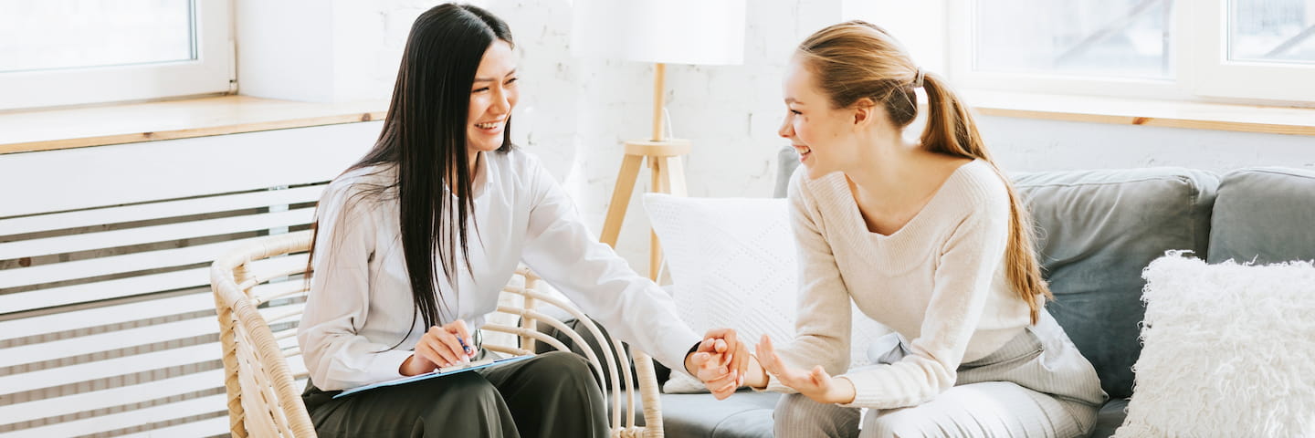Two women smiling and discussing online psychology master's programs while sitting in a bright living room, with one holding a notepad and the other gesturing with her hand.