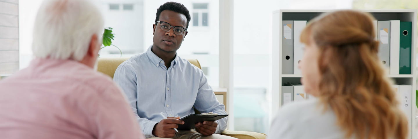 A professional meeting scene with a young black man holding a clipboard while attentively listening across the table to an older couple in a bright, modern office discussing psychology degrees.