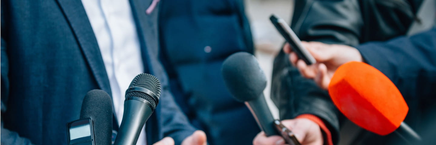 A group of journalists, some likely pursuing online public relations degrees, holding microphones towards a man in a suit during an interview, focusing on the microphones and the man's hands.