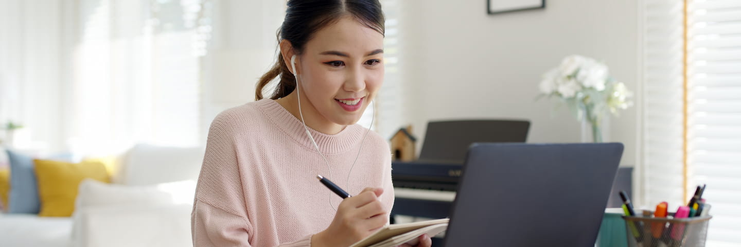 A young woman in a pink sweater, smiling as she writes in her college planning notebook, sitting at a desk with her laptop open and various stationery items around, in a brightly lit room.