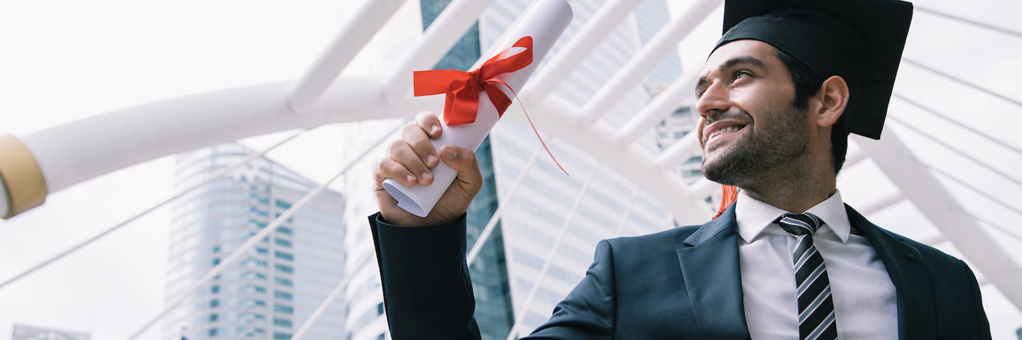 A proud male graduate in a cap and gown, holding his debt-free diploma with a red ribbon, smiles optimistically, while standing outdoors with modern buildings in the background.