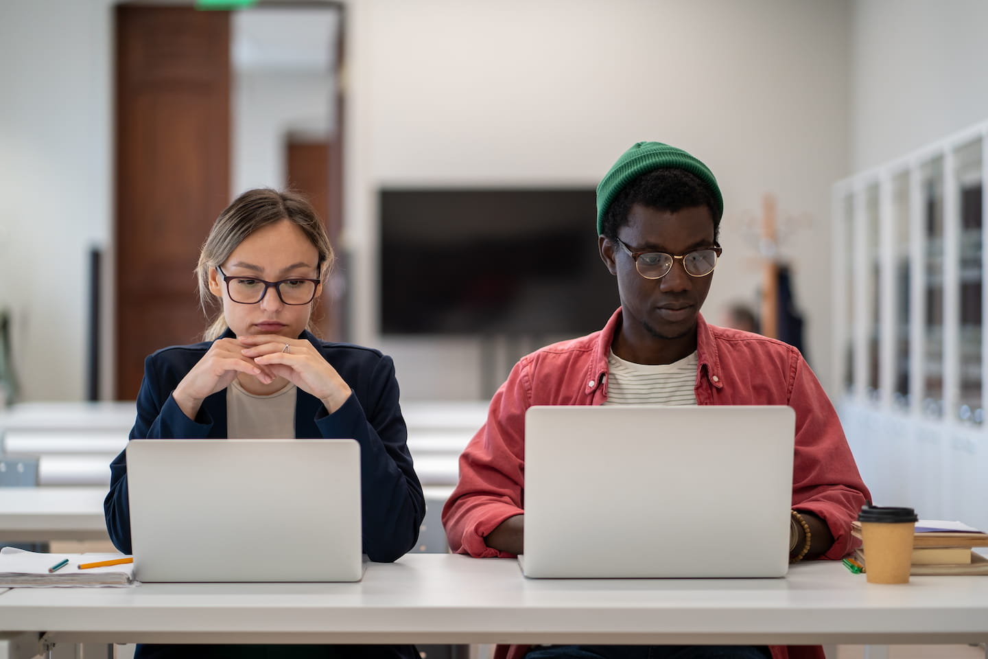 Two young adults, a woman on the left and a man on the right, sit at a table using laptops in a modern office setting. The man wears a green beanie and glasses, both look focused on their screens as they explore different college learning formats.