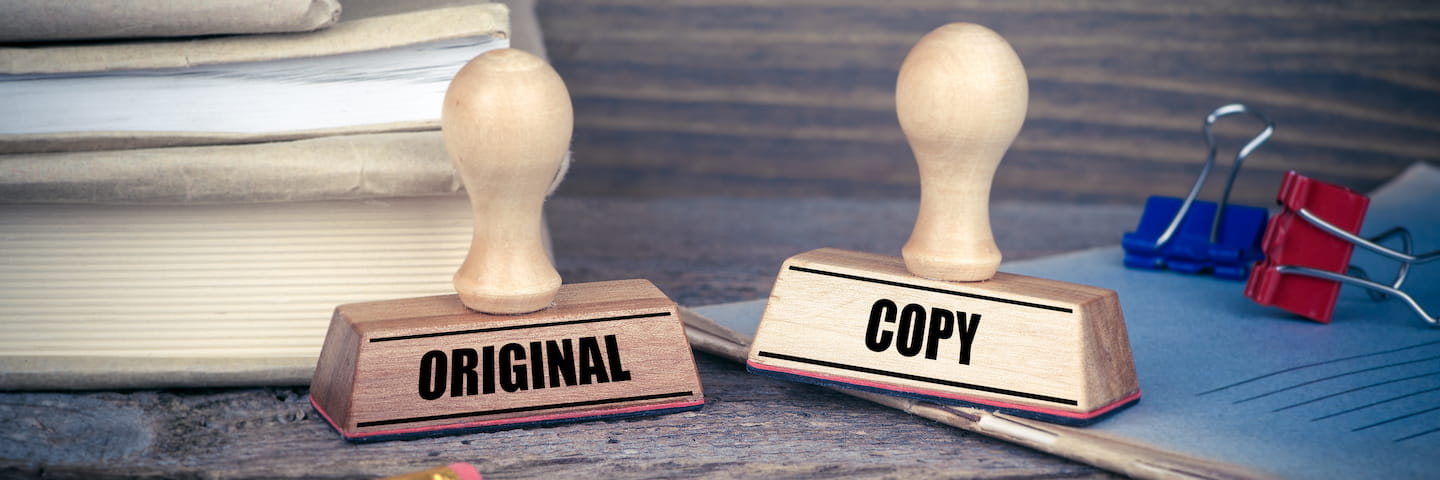 Two wooden rubber stamps on a desk, labeled "original" and "plagiarism" respectively, next to a stack of books and some paperwork with paper clips.