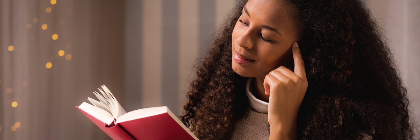 A woman reads a book intently, her finger thoughtfully resting on her cheek. She possesses college-level reading skills and is wearing a cozy sweater, with soft lights in the background. Her curly hair frames her focused expression.