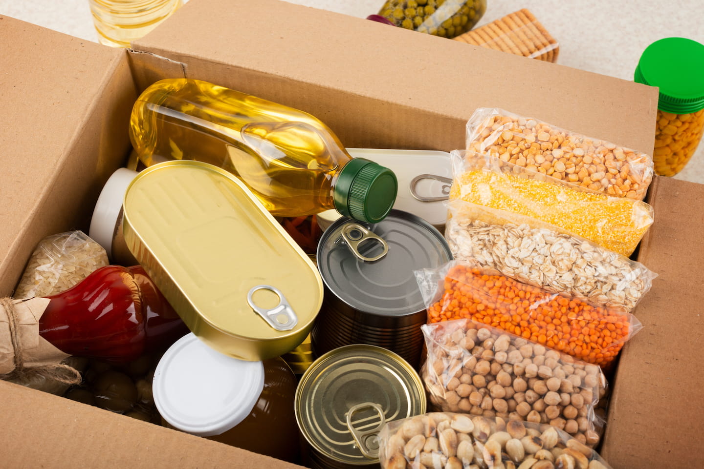 Open cardboard box filled with various non-perishable food items including bottles of oil, dried grains, cans, and jars on a carpeted floor, intended to address student hunger.