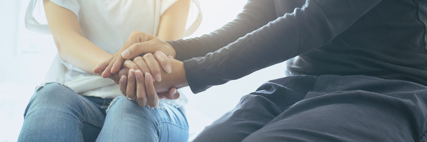 Two college students, sitting opposite each other, holding hands in a comforting manner, illuminated by soft natural light.