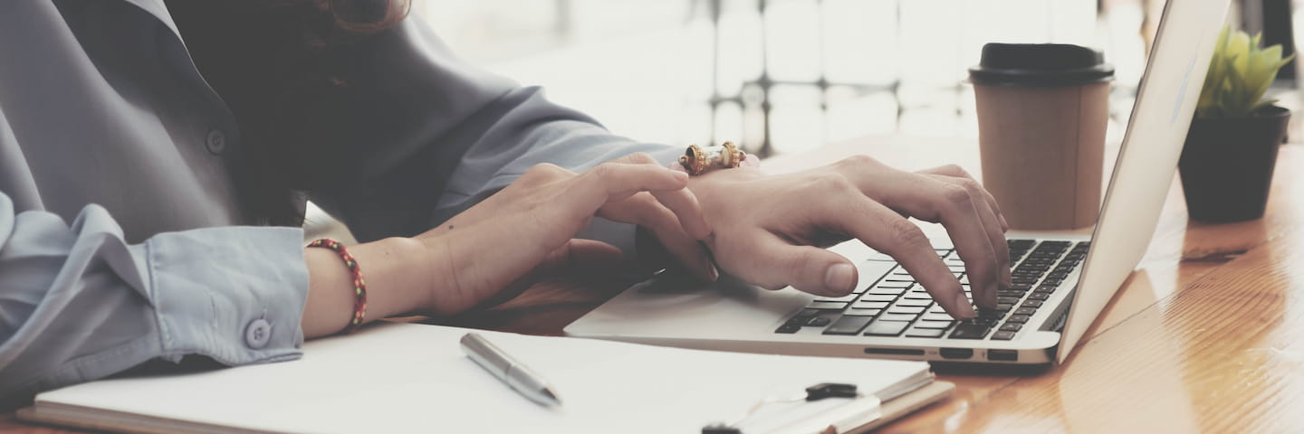A person in a business casual outfit is typing on a laptop at a wooden desk with a disposable coffee cup, college resources, and a pen nearby.