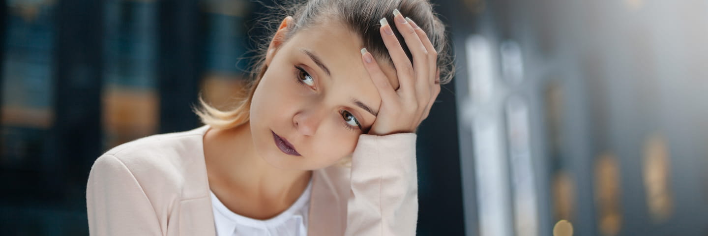 A young college student with blonde hair sitting indoors, resting her head on her hand with a worried expression. She is wearing a lavender blazer over a white top.
