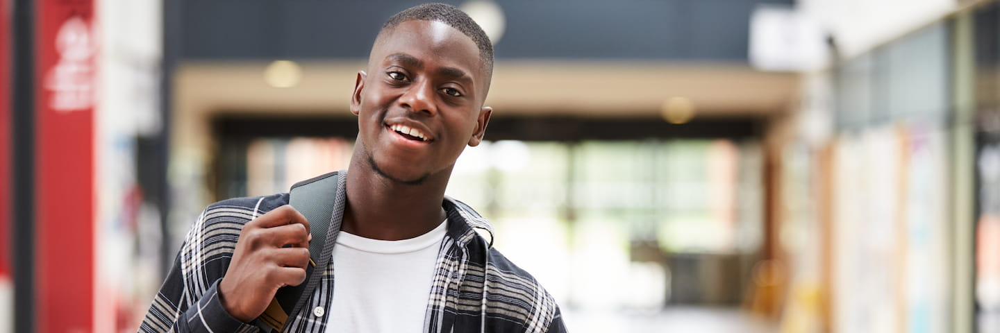 A smiling young man walking in a mall, holding a backpack over one shoulder, wearing a plaid shirt over a white T-shirt as he researches student scholarships.