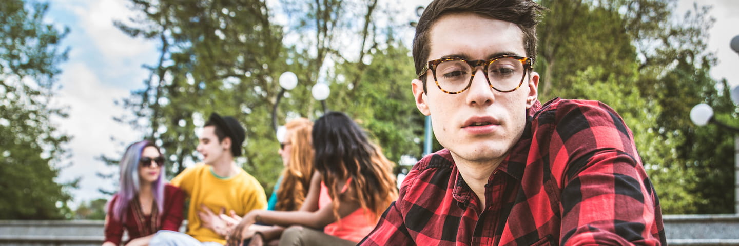 Young man in glasses and a red plaid shirt in the foreground with a group of friends discussing college scholarships in the background in a sunny park setting.
