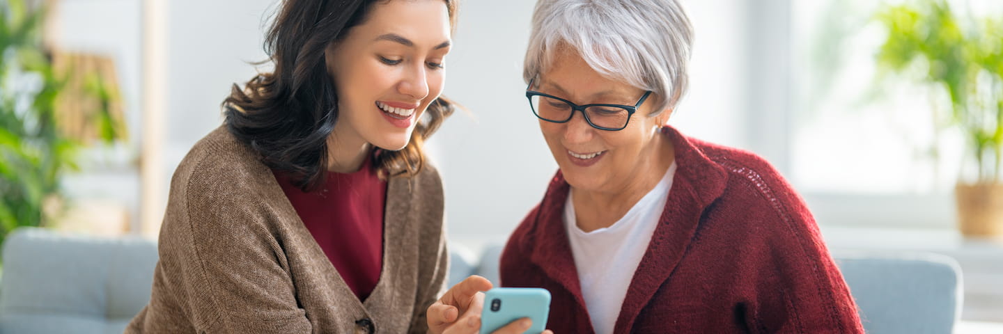 A young woman and an elderly woman smiling and looking at a smartphone together, sitting in a bright, airy room, possibly discussing online DSW programs.