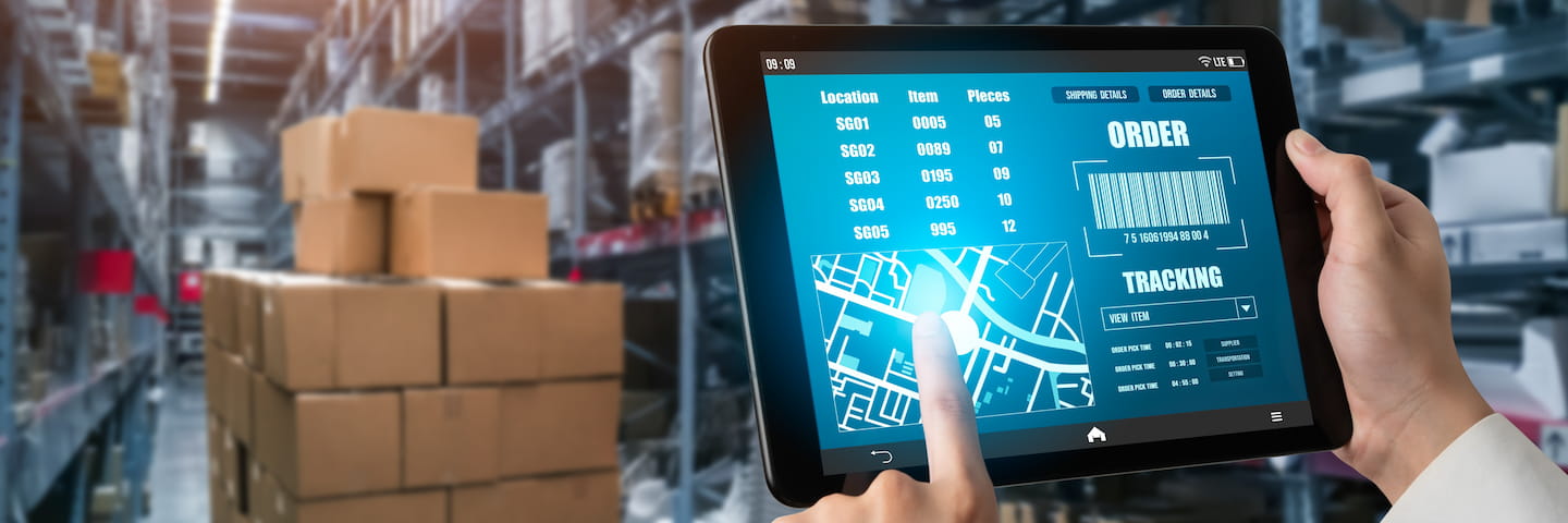 A person in a warehouse holds a tablet showing a supply chain management application with data about inventory location, pieces, and tracking, with blurred shelves of boxes in the background.