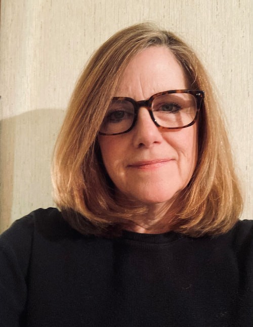 A woman with shoulder-length blond hair and glasses, wearing a black top, smiling slightly at the camera against a textured beige wall background.