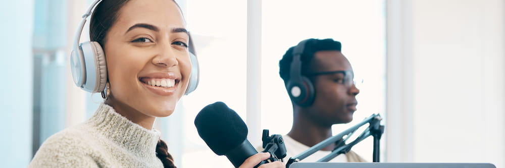 A woman wearing headphones smiles while speaking into a microphone in a radio station studio, with a focused male colleague in the background also wearing headphones.