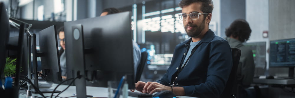 A man with glasses and a beard, wearing a dark shirt, works intently at a computer in a modern office with multiple screens and colleagues nearby.