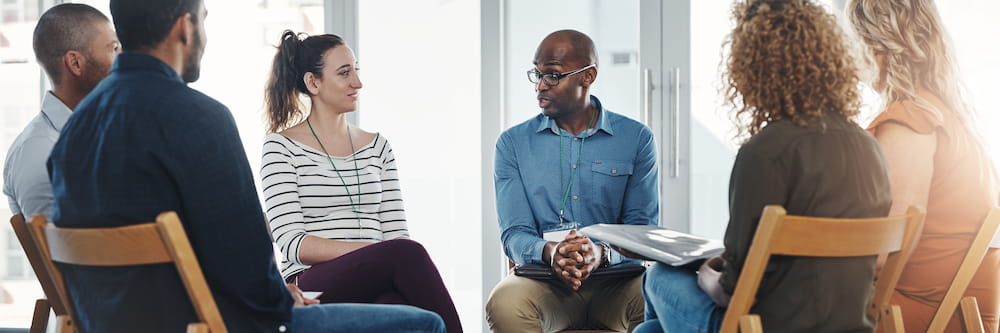 A diverse group of adults sitting in a circle engaging in a discussion, led by a bald Black man wearing glasses, who is speaking to a woman directly across from him.