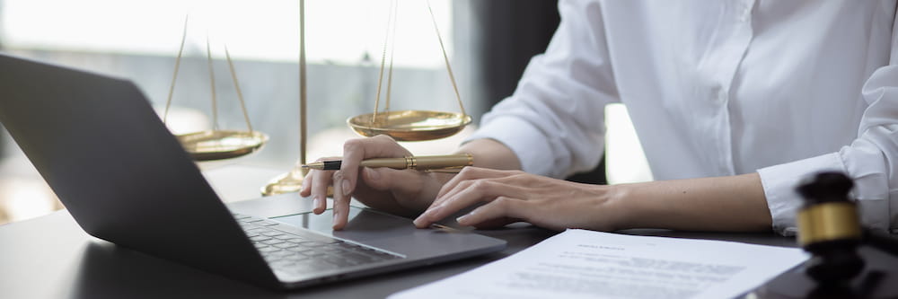 A lawyer in a white shirt works on a laptop in an office, with legal documents, a gavel, and scales of justice on the desk.