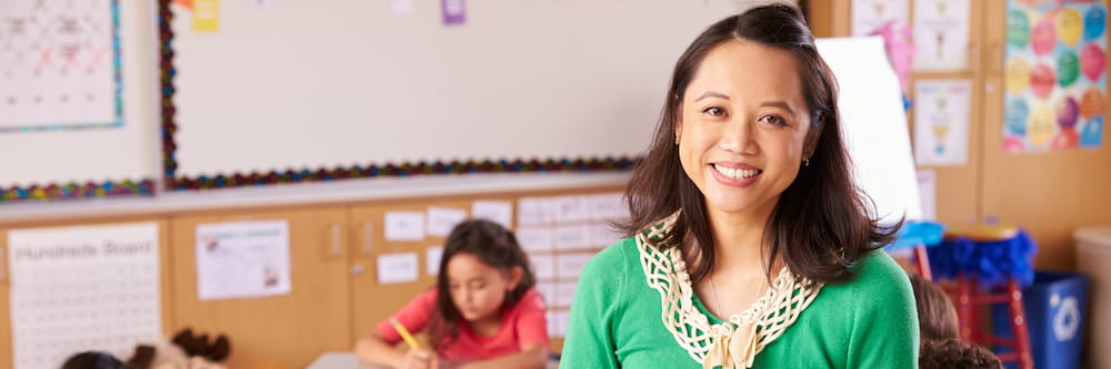 A smiling Asian woman in a green blouse standing in a colorful classroom with children reading books in the background.