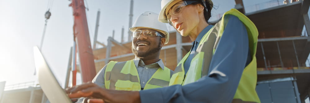 Two construction workers, a man and a woman, wearing hard hats and reflective vests, discuss over a laptop at a construction site during sunset.