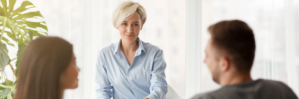 A professional woman with short blonde hair, wearing a blue shirt, smiling and conversing with a couple seated opposite her in a bright, plant-adorned room.