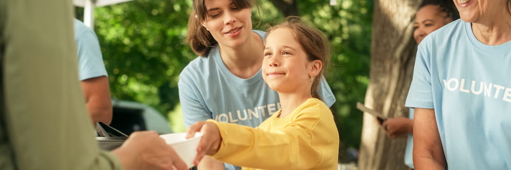 A young girl in a yellow shirt smiles as she receives food from a volunteer, surrounded by other volunteers in blue t-shirts labeled "VOLUNTEER.