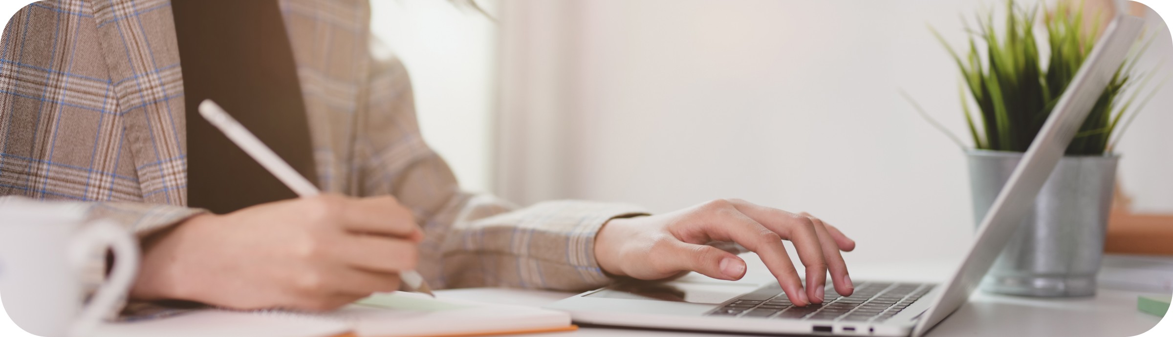 A person in a plaid blazer working on a laptop while writing notes with a potted plant and cup nearby, illustrating a professional office setting for experts.