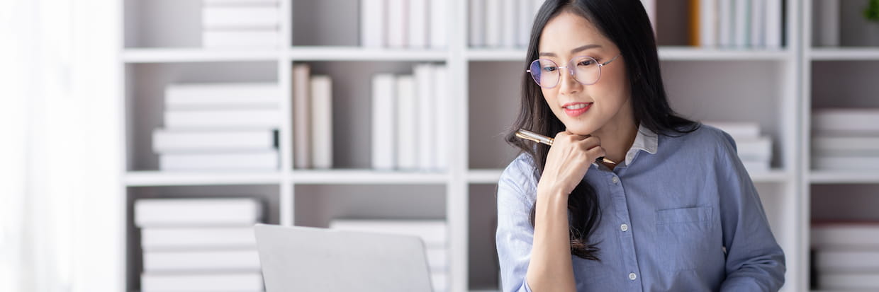 A thoughtful woman with glasses, holding a pen to her chin, sits at a laptop in a library with white shelves full of books, studying to become a Certified Public Accountant.